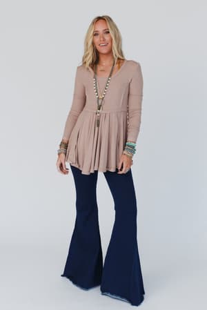A woman with loose blonde hair and a relaxed expression wears The Signature Flare - Dark Denim jeans, a beige peplum top, and layered accessories against a light backdrop.