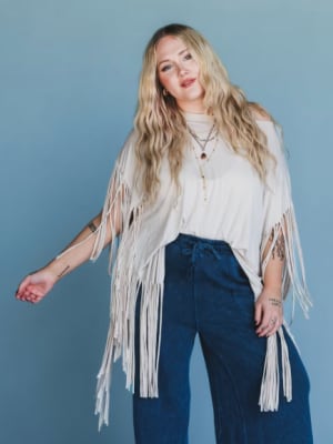 Woman wearing a white fringe cardigan over a white top and blue pants against a blue background