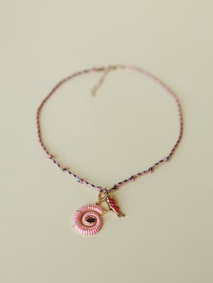 A braided multicolor cord necklace laid flat, featuring a pink enamel spiral shell pendant with a deep red gemstone center and a small gold koi fish charm alongside it — playful and personal, like something you'd pick up on a trip and never take off.
