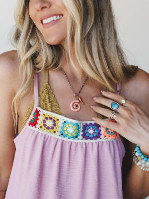 A close-up lifestyle shot capturing a smiling model in a lavender crochet-trim tank, layered with a braided cord necklace featuring a pink shell pendant — the whole look feels like a summer afternoon that stretched into evening.