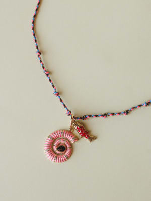 A braided friendship-cord necklace in pink, blue, and black with a gold-toned pink enamel sunburst medallion pendant and a small red enamel koi fish charm — playful, personal, and the kind of piece you never take off.