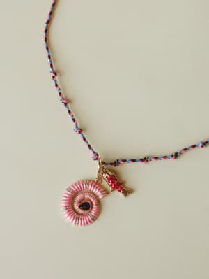 A braided friendship-cord necklace in pink, blue, and black with a gold-toned pink enamel sunburst medallion pendant and a small red enamel koi fish charm — playful, personal, and the kind of piece you never take off.