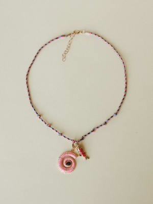 A braided multicolor cord necklace laid flat against a warm neutral background, centered by a pink enamel spiral shell pendant with rhinestone detailing and a small red fish charm — playful, collected, and full of personality.