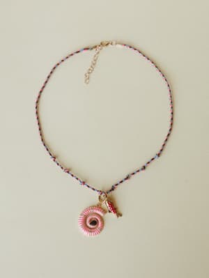A braided multicolor cord necklace laid flat against a warm neutral background, centered by a pink enamel spiral shell pendant with rhinestone detailing and a small red fish charm — playful, collected, and full of personality.