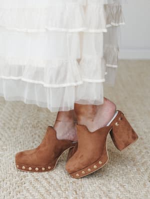 A close-up of caramel suede platform mules with gold stud trim at the sole, styled against the hem of a tiered cream tulle skirt — the kind of pairing that feels effortlessly vintage and a little dreamy.