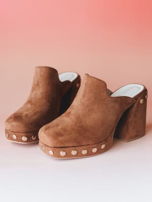 A pair of caramel suede platform clogs shot against a warm blush backdrop — the kind of shoe that makes you want to wear a midi skirt and go somewhere worth walking to.