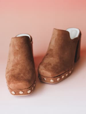 A pair of caramel suede platform clogs shot close-up against a soft blush backdrop — the kind of shoes that make a whole outfit feel intentional without trying too hard.