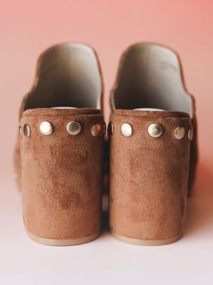 A close-up rear view of caramel suede platform mules with silver stud detailing along the strap — shot against a soft pink backdrop that makes the warm brown leather feel even more cozy and intentional.
