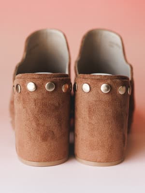 A close-up rear view of caramel suede platform mules with silver stud detailing along the strap — shot against a soft pink backdrop that makes the warm brown leather feel even more cozy and intentional.