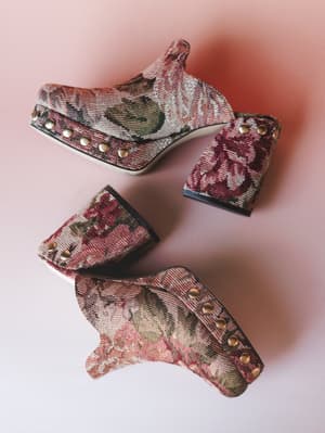 A pair of tapestry platform mules laid casually on a blush pink surface, their floral woven fabric and gold stud trim giving off serious vintage-find energy.