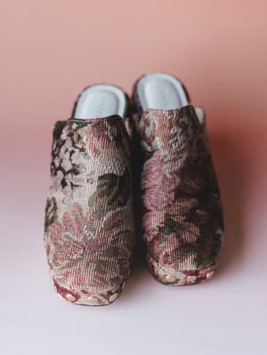 A pair of tapestry-fabric platform mules shot front-facing against a soft blush backdrop — the kind of shoe that looks like it escaped from a vintage garden painting and landed in your closet.
