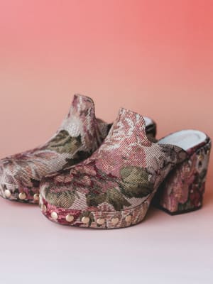 A pair of tapestry-covered platform clogs shot against a warm coral backdrop — the kind of shoes that make a whole outfit feel intentional without trying too hard.