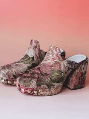 A pair of tapestry-covered platform clogs shot against a warm coral backdrop — the kind of shoes that make a whole outfit feel intentional without trying too hard.