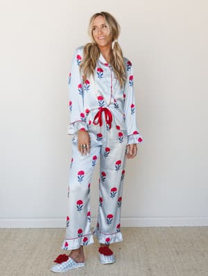A full-length satin pajama set in pale blue with a red floral block print — the kind of set that makes staying in feel like a whole personality, styled with gingham slippers and dangling pearl earrings.