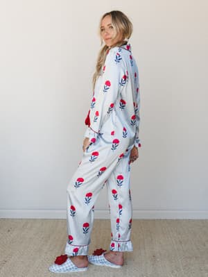 A relaxed full-body shot of a model in a matching floral pajama set — long-sleeve button-up top and wide-leg pants in soft white with red blooms and navy stems — styled with gingham slippers and pearl earrings, glancing back over her shoulder with ea