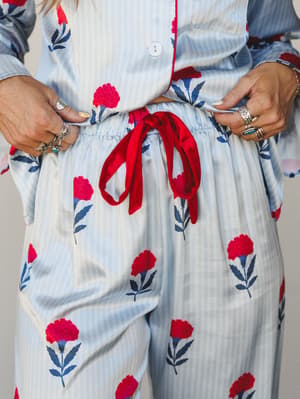 A close-up shot of a matching floral pajama set in soft blue pinstripe with bold red carnation blooms and navy leaf stems — styled with a stack of silver rings and a red satin drawstring bow that makes it feel almost too pretty to sleep in.
