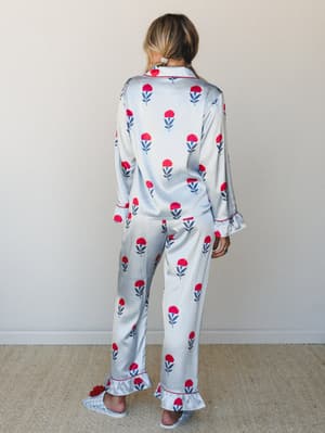 A satin floral pajama set shot from the back — the kind of set that makes staying in feel like an occasion, with red carnation blooms on pale blue stripe, ruffle hem details, and matching floral slippers.