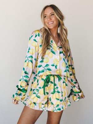 A lemon-print satin two-piece set — cropped button-up top and ruffle-hem shorts — worn with effortless summer energy against a clean neutral backdrop.