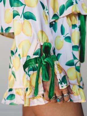 A close-up detail shot of a lemon-print PJ short set with ruffle trim and a velvet green tie closure at the waist, styled with a stack of silver and turquoise rings — the kind of set that makes staying in feel intentional.