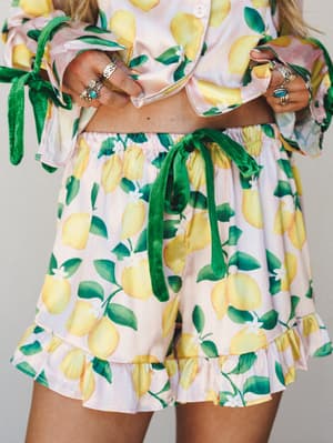 A close-up of a lemon-print matching set — cropped top and ruffled shorts — styled with stacked silver rings and green velvet tie details that give the whole look a playful, summery sweetness.