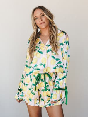 A matching lemon-print satin pajama set — button-front top and ruffle-hem shorts with a green satin tie — styled on a model against a soft neutral studio wall with an easy, sun-drenched summer feel.