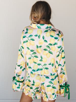 A model shot from behind in a satin lemon-print pajama set — the long-sleeve top and ruffled shorts feel equal parts vacation fantasy and Sunday morning energy.