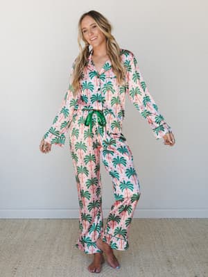 A satin palm tree print pajama set — button-front top and wide-leg pants — worn with easy confidence against a clean studio wall, the kind of set that makes staying in feel like a whole occasion.