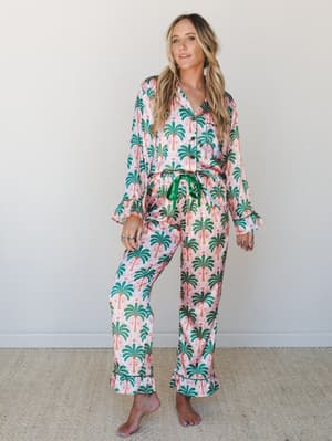 A matching satin pajama set in a bold palm tree print — blush pink with green and orange — styled with a green drawstring tie and ruffle cuffs that make it feel more 'vacation morning' than 'actually sleeping.'