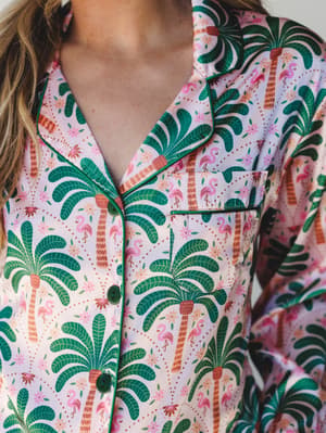 A close-up of a silky button-front pajama top covered in a bold tropical print — palm trees, pink flamingos, and tiny florals — finished with deep green contrast piping and matching buttons that make the whole thing feel more like a vacation souvenir