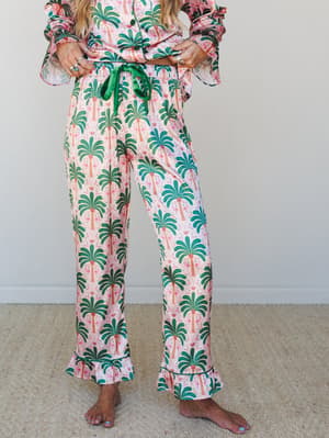 A palm tree and flamingo printed matching PJ set in blush pink and emerald green, styled on a barefoot model from the waist down — playful, relaxed, and the kind of set you'd actually wear all day.