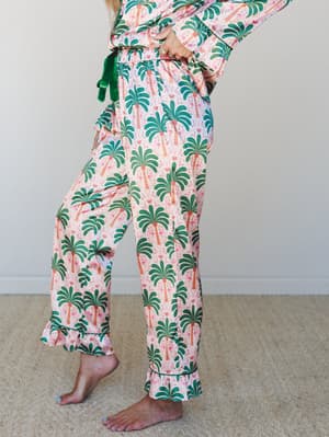 A side-angle shot of wide-leg pajama pants in a playful pink, green, and orange palm tree and flamingo print — ruffle-hemmed with a green velvet drawstring tie, worn barefoot indoors for that effortlessly cozy, resort-at-home feel.