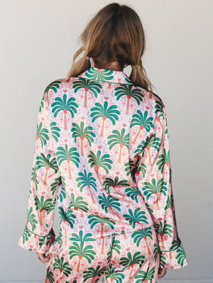 A back-facing shot of a satin pajama set covered in a bold palm tree and flamingo print — the kind of set that makes you want to call it 'resort wear' and mean it.