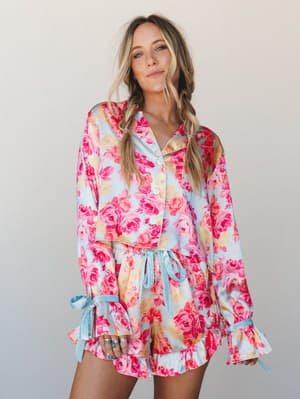 A floral satin PJ short set in soft blue and hot pink roses, styled with braided pigtails and stacked rings — the kind of set that makes getting dressed feel like a whole moment.
