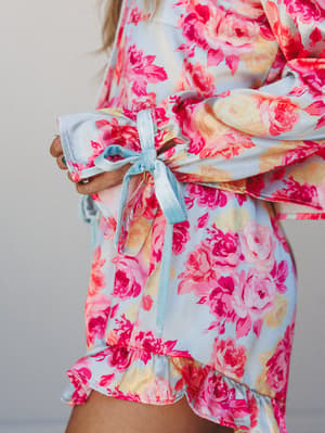 A close-up of a satin floral PJ short set in powder blue and hot pink roses, tied at the waist with a soft ribbon bow — the kind of thing you wear on a slow morning and refuse to take off.