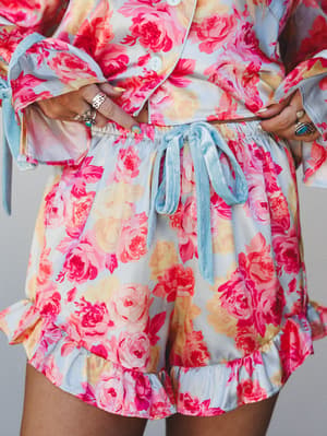 A close-up mid-shot of a floral two-piece short set in soft pink and sky blue, styled with stacked silver and turquoise rings — the kind of set that blurs the line between pajamas and something you'd actually wear to brunch.