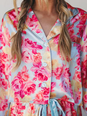 A close-up of a satin floral pajama top in powder blue and hot pink roses, buttoned up with a notch collar and a tie detail at the hem — the kind of set that makes staying in feel like a whole moment.