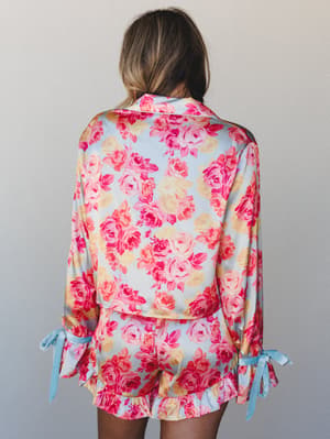 A floral satin pajama set shot from the back — the oversized top and ruffle-hem shorts share a dreamy rose print in pink and periwinkle, finished with soft bow-tied cuffs that feel equal parts vintage and sweet.