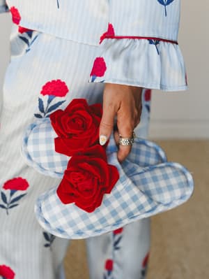 A close-up of a hand holding blue gingham slippers adorned with velvet red roses, styled against a coordinating floral-print pajama set — the kind of cozy detail that makes staying in feel just as intentional as going out.