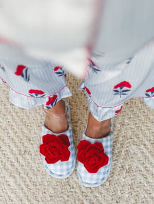 A top-down shot of gingham slippers with bold red rose embellishments, peeking out from under the hem of a floral-printed striped skirt — the kind of detail you'd photograph yourself wearing on a slow morning.