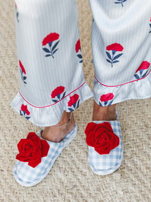 A close-up of wide-leg pajama pants with a red floral and navy stripe print, paired with blue gingham slippers topped with oversized red rosette appliqués — the kind of cozy detail that makes staying in feel like a whole look.