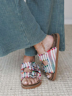 A close-up of colorful fringe-trimmed slide sandals with silver buckle hardware, worn with a flowy teal gauze wide-leg pant and a delicate gold anklet — the kind of detail shot that makes you want to look down at your own feet.