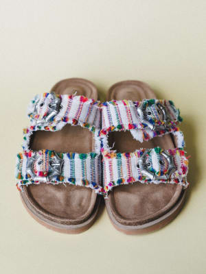 A pair of double-strap slide sandals shot overhead against a soft sage background — the woven fabric straps are bursting with multicolor fringe and finished with silver hardware, sitting on a worn-in suede footbed that feels instantly comfortable.
