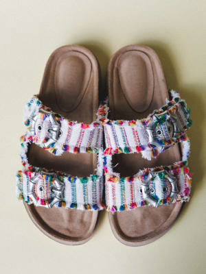 A pair of double-strap slide sandals shot overhead on a soft sage background — the fringe-edged woven straps burst with multicolor threads, grounded by silver star buckles and a warm tan footbed.