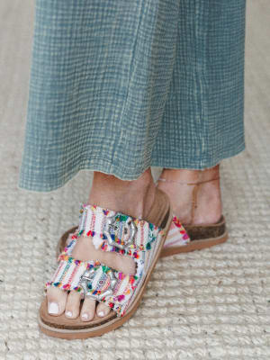 A close-up of colorful frayed fabric slide sandals with silver medallion buckle hardware, paired with a breezy sage gauze wide-leg pant — the kind of shoe that does all the talking.