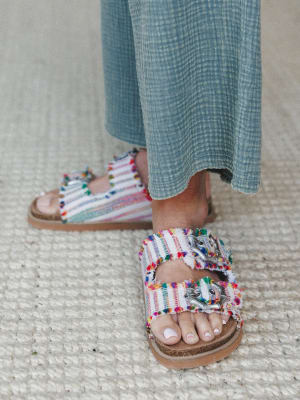 A close-up of colorful fringe-edged slide sandals with silver buckle hardware, worn with a breezy blue gauze skirt — the kind of shoes that make a simple outfit feel like a whole personality.