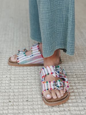 A close-up of colorful fringe-edged slide sandals with silver buckle hardware, worn with a breezy blue gauze skirt — the kind of shoes that make a simple outfit feel like a whole personality.