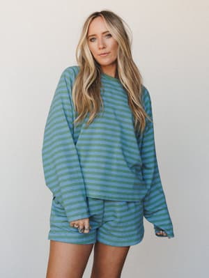 A matching striped sweatshirt-and-shorts set in sage green and periwinkle blue, worn relaxed and easy on a neutral studio backdrop — the kind of outfit that makes staying in feel like a whole personality.