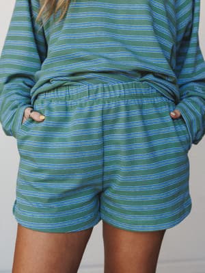 A close-up shot of a matching striped short set in sage green and blue — hands tucked into pockets, relaxed fit, the kind of outfit that makes staying home feel like a whole personality.