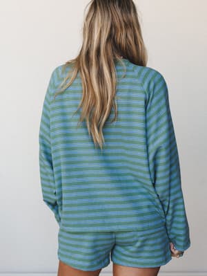 Shot from behind, a model wears a matching striped pullover and shorts set in sage green and blue — the kind of easygoing set that blurs the line between staying in and stepping out.