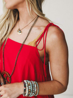 A close-up lifestyle shot of a red tie-shoulder dress styled with layered gold and chain necklaces, a stack of boho beaded bracelets, and a peeking lace bralette — the kind of outfit that looks effortless but definitely wasn't accidental.